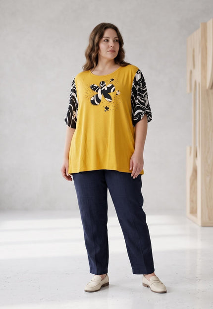 Ολόσωμη λήψη με γυναικεία κίτρινη τουνίκ με σχέδιο και ασπρόμαυρα μανίκια, σε μεγάλα μεγέθη, συνδυασμένη με σκούρο μπλε παντελόνι.