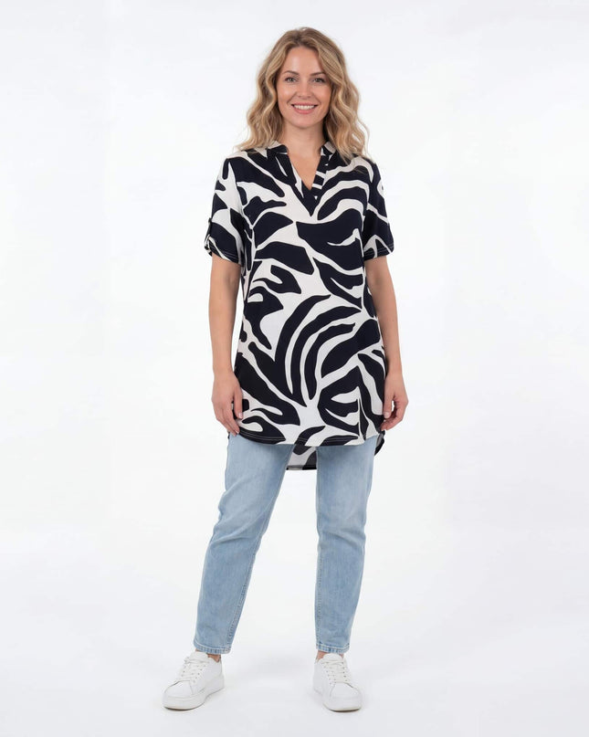 Ολόσωμη λήψη: Γυναίκα φοράει ασύμμετρη ασπρόμαυρη καλοκαιρινή τουνίκ σε μεγάλα μεγέθη με αφηρημένο σχέδιο, συνδυασμένη με τζιν.