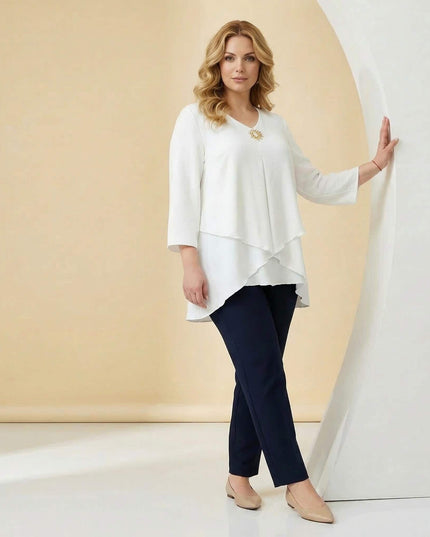 Ολόσωμη λήψη μοντέλου που φοράει επίσημη λευκή τουνίκ σε μεγάλα μεγέθη, με ασύμμετρη εβαζέ γραμμή και χρυσή καρφίτσα στο ντεκολτέ.