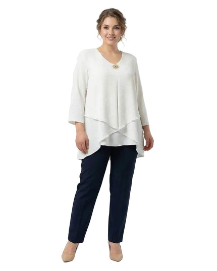 Ολόσωμη λήψη σε λευκό φόντο. Γυναίκα με επίσημη λευκή τουνίκ σε μεγάλα μεγέθη, με ασύμμετρη γραμμή σε επίπεδα, 7/8 μανίκια και χρυσή καρφίτσα.