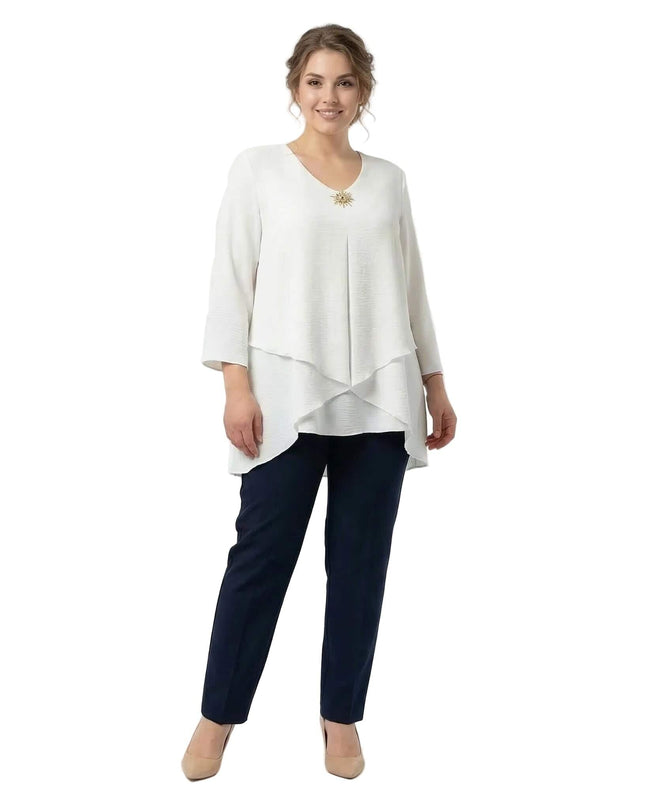 Ολόσωμη λήψη σε λευκό φόντο. Γυναίκα με επίσημη λευκή τουνίκ σε μεγάλα μεγέθη, με ασύμμετρη γραμμή σε επίπεδα, 7/8 μανίκια και χρυσή καρφίτσα.