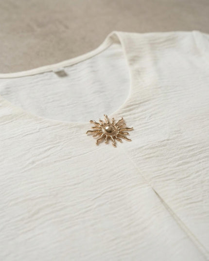 Κοντινή λεπτομέρεια στο V ντεκολτέ από επίσημη λευκή τουνίκ μεγάλα μεγέθη, που δείχνει τη χρυσή καρφίτσα ήλιο και την ανάγλυφη υφή του υφάσματος.