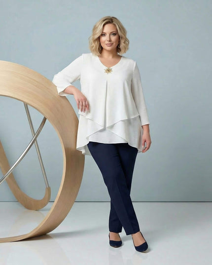 Ολόσωμη λήψη γυναίκας που φοράει επίσημη λευκή τουνίκ σε μεγάλα μεγέθη. Ξεχωρίζει η ασύμμετρη γραμμή σε επίπεδα, τα μανίκια 7/8 και η χρυσή καρφίτσα.