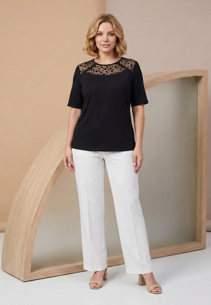 Ολόσωμη λήψη επίσημης μαύρης μπλούζας plus size με εντυπωσιακό ντεκολτέ από τούλι και λευκό παντελόνι.