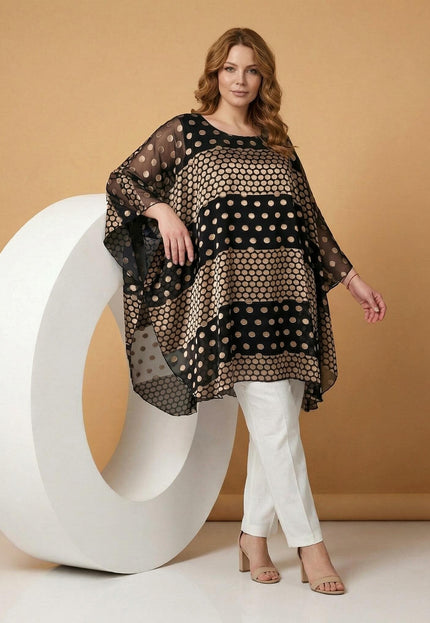 Ολόσωμη λήψη. Επίσημη μαύρη plus size τουνίκ πόντσο με εντυπωσιακά χρυσά πουά σε διαφανές σιφόν. Το μοντέλο ποζάρει με λευκό παντελόνι.