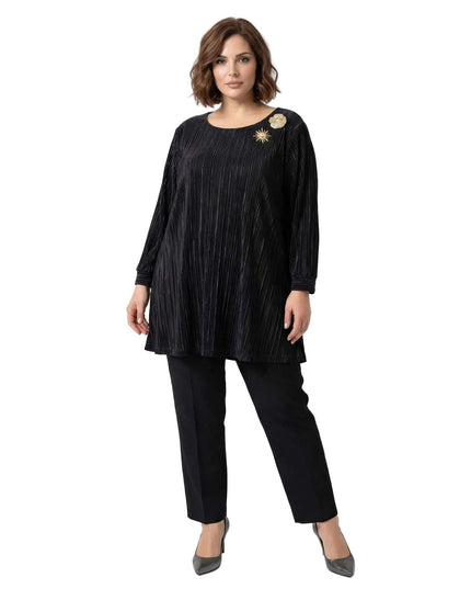 Ολόσωμη λήψη με μοντέλο που φοράει κομψή μαύρη τουνίκ σε μεγάλα μεγέθη, κατασκευασμένη από πλισέ ύφασμα. Ξεχωρίζει η χρυσή καρφίτσα στο ύψος του ώμου.