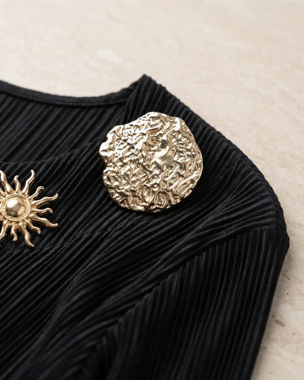 Κοντινή λεπτομέρεια από μαύρη τουνίκ σε μεγάλα μεγέθη, με εστίαση στην πλισέ υφή του υφάσματος και τις εντυπωσιακές χρυσές καρφίτσες στο πάνω μέρος.