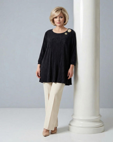 Ολόσωμη λήψη της επίσημης μαύρης τουνίκ σε μεγάλα μεγέθη από πλισέ σολεΐ με χρυσή καρφίτσα, φορεμένη από μοντέλο δίπλα σε λευκή κολώνα.