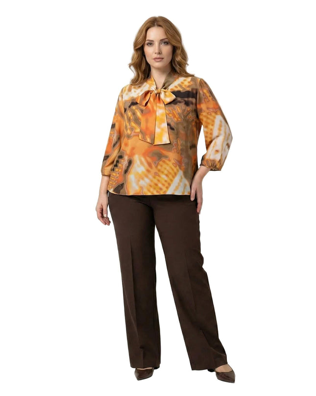 Επίσημη πορτοκαλί τουνίκ plus size με αφηρημένο σχέδιο, φιόγκο στο λαιμό και λάστιχο στο κάτω μέρος, σε ολόσωμη λήψη με καφέ παντελόνι.