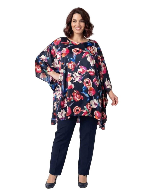 Ολόσωμη λήψη γυναίκας που φοράει επίσημη σκούρο μπλε φλοράλ τουνίκ πόντσο για μεγάλα μεγέθη, με ασύμμετρο τελείωμα και σκούρο μπλε παντελόνι.