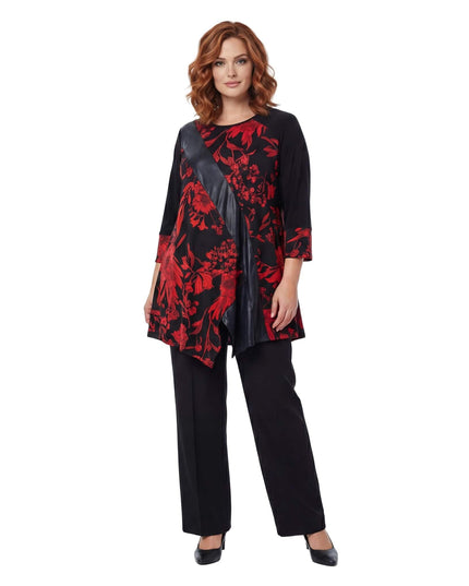 Επίσημη γυναικεία τουνίκ σε μεγάλα μεγέθη με κόκκινα λουλούδια και διαγώνια φάσα από δερματίνη, σε ολόσωμη λήψη με μαύρο παντελόνι.