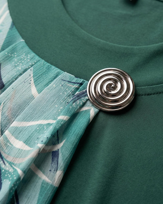 Κοντινή λεπτομέρεια από επίσημη τουνίκ μεγάλα μεγέθη σε λαδί χρώμα, εστιάζοντας στην ασημένια καρφίτσα σπιράλ που συγκρατεί το πτυχωτό φλοράλ σιφόν ύφασμα.