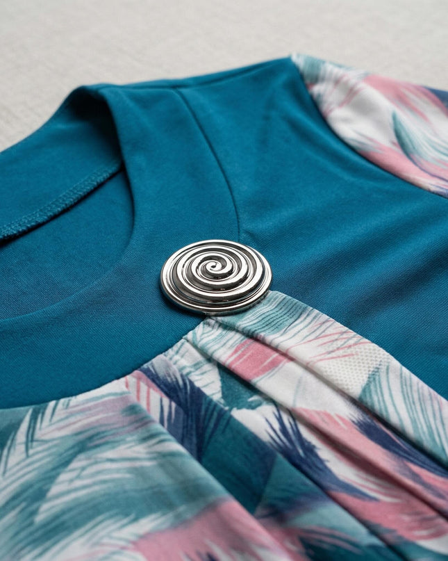 Κοντινή λεπτομέρεια από επίσημη τουνίκ πετρόλ μεγάλα μεγέθη, που εστιάζει στην ασημένια σπιράλ καρφίτσα στο ντεκολτέ και τις πτυχώσεις του εμπριμέ υφάσματος.