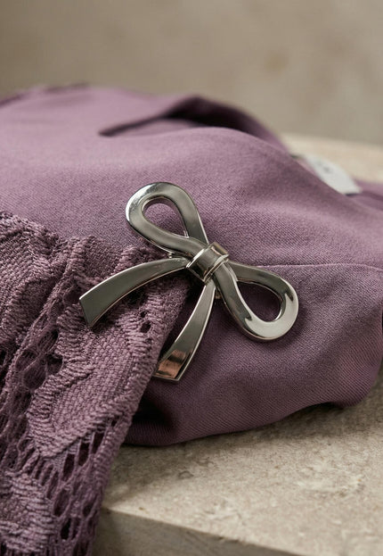 Κοντινή λεπτομέρεια από επίσημη τουνίκ plus size σε σάπιο μήλο, με εστίαση στη λαμπερή μεταλλική καρφίτσα σε σχήμα φιόγκου και στο ύφασμα με δαντέλα.