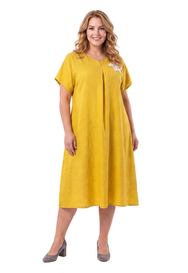 Κίτρινο plus size εβαζέ φόρεμα με V λαιμόκοψη, μπροστινή πιέτα και λευκή φλοράλ απλικέ λεπτομέρεια στον ώμο, σε λευκό φόντο.