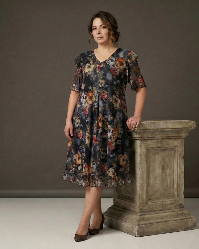 Επίσημο φλοράλ φόρεμα σιφόν σε μεγάλα μεγέθη, με αέρινα μανίκια και βολάν στο τελείωμα. Ολόσωμη λήψη μοντέλου που στέκεται δίπλα σε πέτρινο βάθρο.