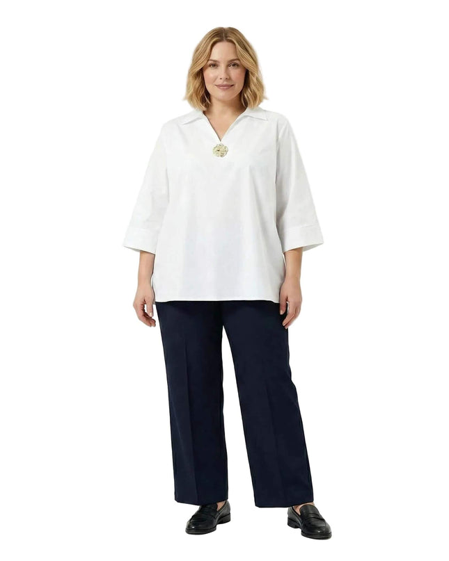 Ολόσωμη λήψη με γυναικείο επίσημο λευκό βαμβακερό πουκάμισο σε μεγάλα μεγέθη, διακοσμημένο με καρφίτσα στο ντεκολτέ και συνδυασμένο με σκούρο παντελόνι.