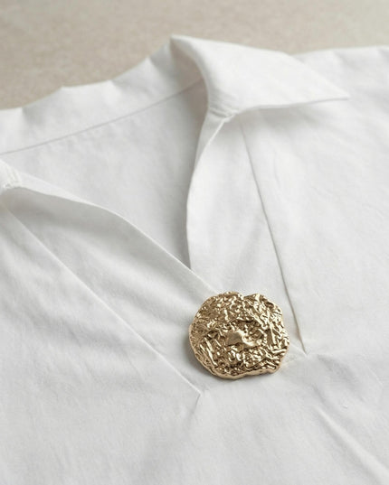 Κοντινή λεπτομέρεια από επίσημο λευκό βαμβακερό πουκάμισο σε μεγάλα μεγέθη, με εστίαση στο ντεκολτέ και τη διακοσμητική χρυσή καρφίτσα.