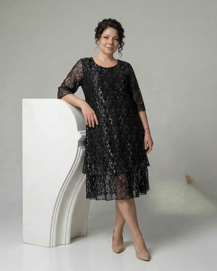 Ολόσωμη λήψη, επίσημο μαύρο φόρεμα σε μεγάλα μεγέθη με εντυπωσιακή δαντέλα, 3/4 μανίκια και κλιμακωτό τελείωμα. Το μοντέλο σε λευκό φόντο.