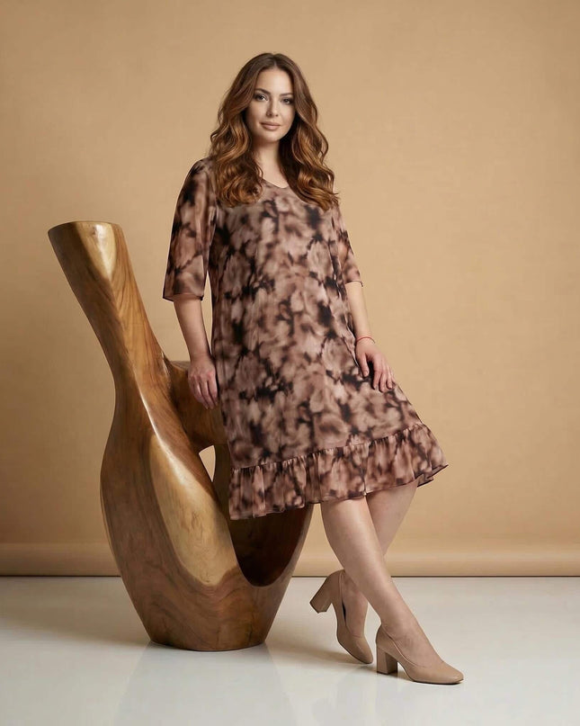 Ολόσωμη λήψη γυναίκας που ποζάρει δίπλα σε ξύλινο γλυπτό, φορώντας ένα επίσημο μάξι φόρεμα μεγάλα μεγέθη από αέρινο σιφόν με ντεγκραντέ σχέδιο.