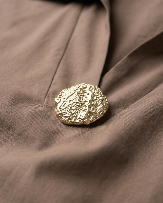 Κοντινή λεπτομέρεια του χρυσού διακοσμητικού στοιχείου στο ντεκολτέ από το επίσημο βαμβακερό πουκάμισο μόκα, σε άνετη γραμμή για plus size σωματότυπους.