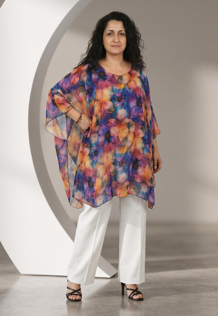 Άτομο φορά πολύχρωμη φλοράλ τουνίκ τύπου πόντσο με φαρδιά μανίκια και μαύρο περίγραμμα, πάνω από λευκό παντελόνι.
