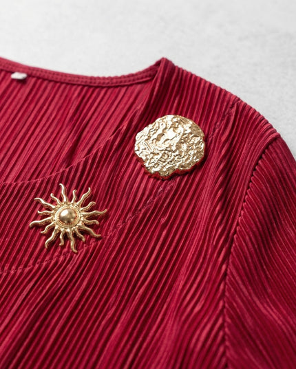 Κοντινή λεπτομέρεια από την κόκκινη τουνίκ σε μεγάλα μεγέθη, όπου διακρίνονται το πλισέ ύφασμα και οι δύο χρυσές καρφίτσες.