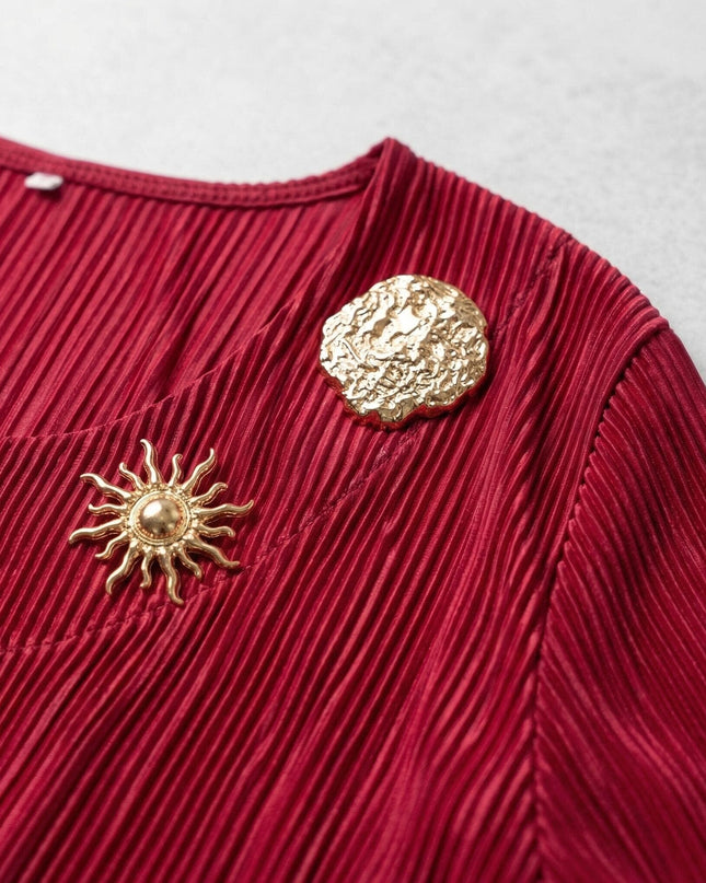 Κοντινή λεπτομέρεια από την κόκκινη τουνίκ σε μεγάλα μεγέθη, όπου διακρίνονται το πλισέ ύφασμα και οι δύο χρυσές καρφίτσες.