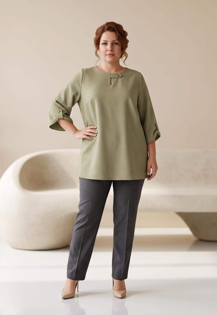 Ολόσωμη λήψη με γυναίκα plus size που φοράει κομψή επίσημη τουνίκ σε λαδί χρώμα με μανίκια 3/4, συνδυασμένη με γκρι παντελόνι.