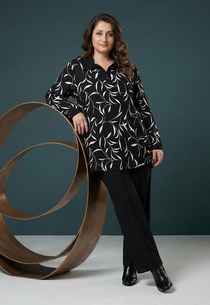Κομψή γυναικεία τουνίκ plus size με αφηρημένο print και μαύρο γιακά, σε ολόσωμη λήψη με το μοντέλο να ακουμπά σε μεταλλικό διακοσμητικό.