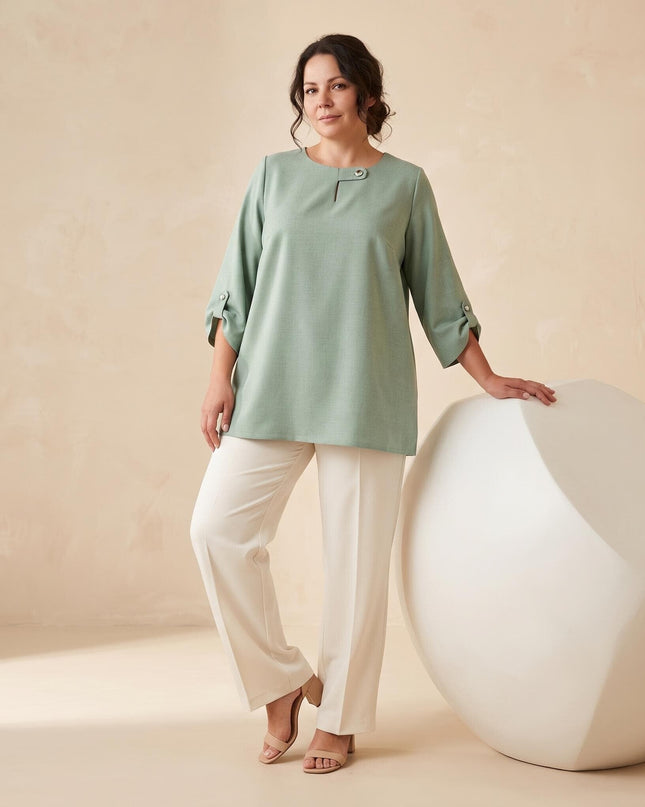 Ολόσωμη λήψη με γυναίκα plus size που φοράει κομψή μακριά τουνίκ σε απαλό πράσινο χρώμα, συνδυασμένη με λευκό παντελόνι.