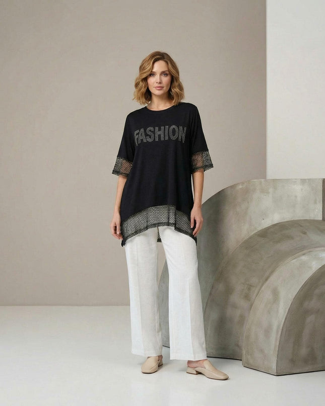 Ολόσωμη λήψη: Κομψή μαύρη ασύμμετρη τουνίκ plus size με δίχτυ στα μανίκια και λεπτομέρειες από στρας. Φορεμένη με λευκό παντελόνι.