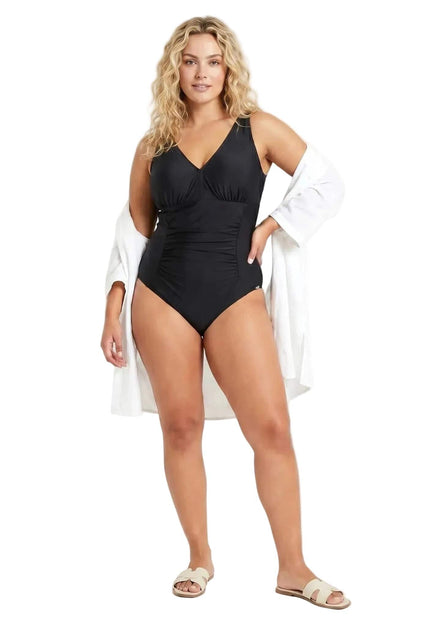 Κομψό μαύρο ολόσωμο μαγιό plus size με V ντεκολτέ και ντραπέ σχέδιο. Ολόσωμη λήψη σε λευκό φόντο με το μοντέλο να φοράει ανοιχτό λευκό πουκάμισο.