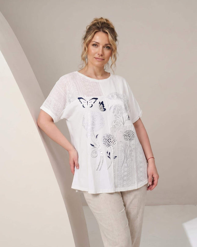 Λευκή βαμβακερή τουνίκ plus size με φλοράλ απλικέ και πάνελ δαντέλας. Μπροστινή όψη σε 3/4 κάδρο, φορεμένη από μοντέλο με ανοιχτόχρωμο παντελόνι.