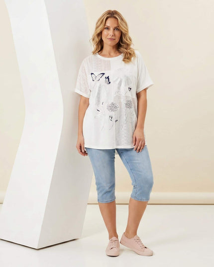 Ολόσωμη λήψη με γυναίκα που φοράει λευκή βαμβακερή τουνίκ plus size με φλοράλ απλικέ και δαντέλα, συνδυασμένη με γαλάζιο κάπρι παντελόνι.