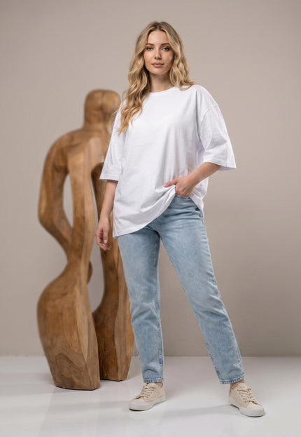Ολόσωμη λήψη: Γυναικείο λευκό βαμβακερό t-shirt σε ίσια γραμμή για μεγάλα μεγέθη, φορεμένο με τζιν, με ξύλινο γλυπτό στο φόντο.