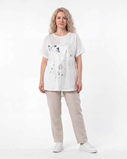 Ολόσωμη λήψη της λευκής βαμβακερής τουνίκ plus size με φλοράλ απλικέ και δαντέλα σε συνδυασμό με μπεζ παντελόνι και λευκά sneakers σε λευκό φόντο.