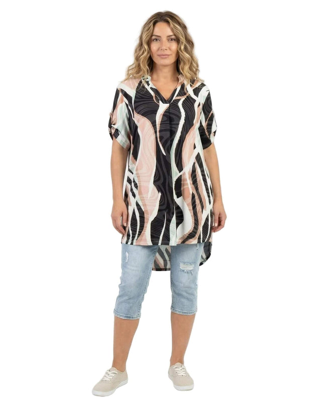 Ολόσωμη λήψη με γυναίκα να φοράει μακριά ασύμμετρη τουνίκ plus size με αφηρημένο πολύχρωμο μοτίβο και V λαιμόκοψη, συνδυασμένη με τζιν.