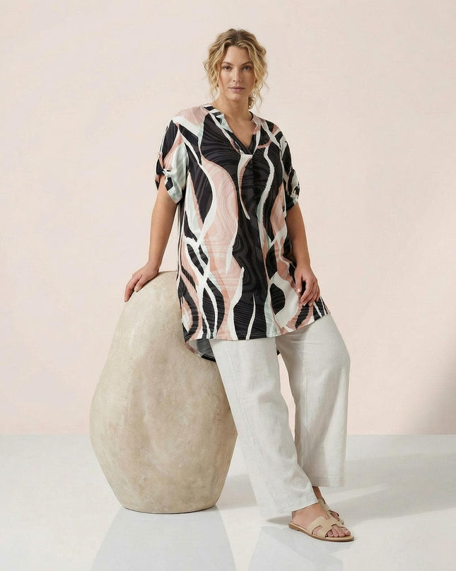 Ολόσωμη λήψη: Γυναίκα με μακριά ασύμμετρη καλοκαιρινή τουνίκ σε μεγάλα μεγέθη, διακοσμημένη με αφηρημένο πολύχρωμο μοτίβο και λαιμόκοψη V.