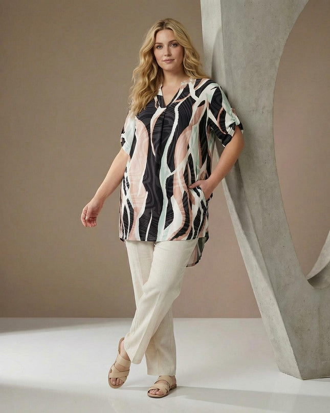 Ολόσωμη λήψη μοντέλου που φοράει μακριά ασύμμετρη τουνίκ plus size με πολύχρωμο κυματιστό μοτίβο, συνδυασμένη με ανοιχτόχρωμο παντελόνι.