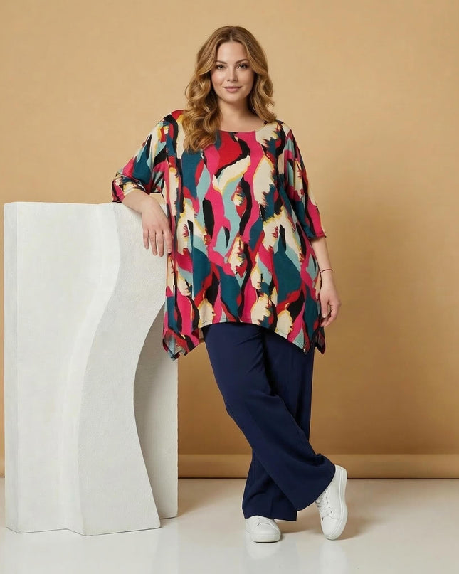 Ολόσωμη λήψη μοντέλου με μακριά καλοκαιρινή plus size τουνίκ σε πολύχρωμο αφηρημένο μοτίβο, συνδυασμένη με σκούρο μπλε παντελόνι.