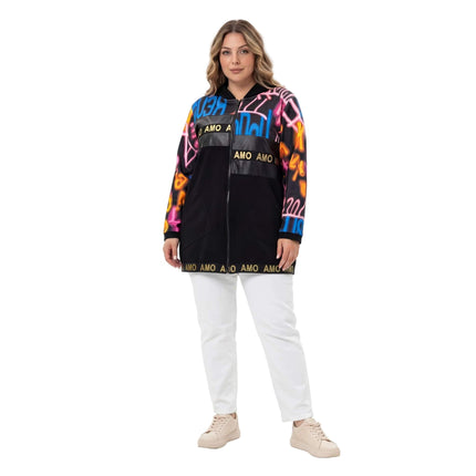Ολόσωμη λήψη, μαύρη γυναικεία ζακέτα plus size με κουκούλα, κεντρικό φερμουάρ και πολύχρωμο τύπωμα γκράφιτι στα μανίκια, φορεμένη με λευκό παντελόνι.