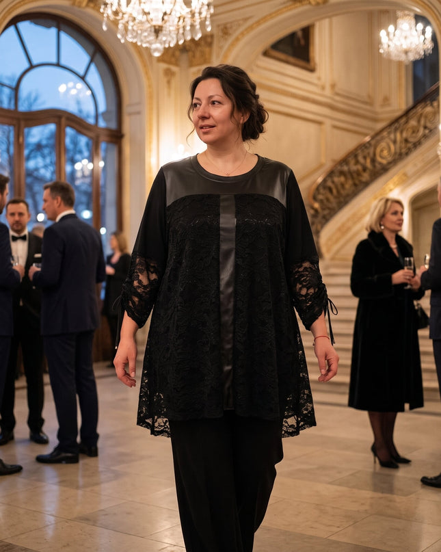 Γυναίκα φορά μαύρη τουνίκ με δαντελένια πάνελ και γυαλιστερή κάθετη λωρίδα μπροστά, σε επίσημη αίθουσα με πολυελαίους και καλεσμένους.