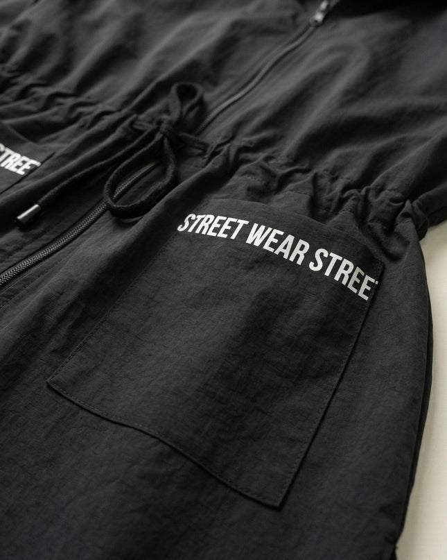 Κοντινή λεπτομέρεια σε μαύρο αντιανεμικό μπουφάν για μεγάλα μεγέθη, που αναδεικνύει το κορδόνι στη μέση και τη λευκή στάμπα Street Wear στην τσέπη.