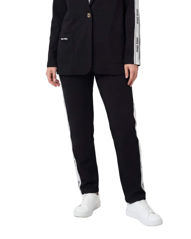 Μαύρο καθημερινό γυναικείο σετ σακάκι και παντελόνι σε μεγάλα μεγέθη, με λευκή διακοσμητική ρίγα στο πλάι και λευκά αθλητικά, σε λευκό φόντο.