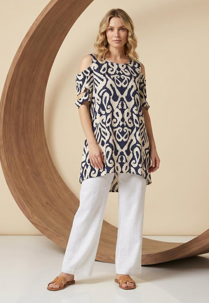 Μπεζ καλοκαιρινή τουνίκ με ανοιχτούς ώμους και έντονο μπλε μοτίβο, ιδανική για μεγάλα μεγέθη. Ολόσωμη λήψη με μοντέλο που φοράει λευκό παντελόνι.