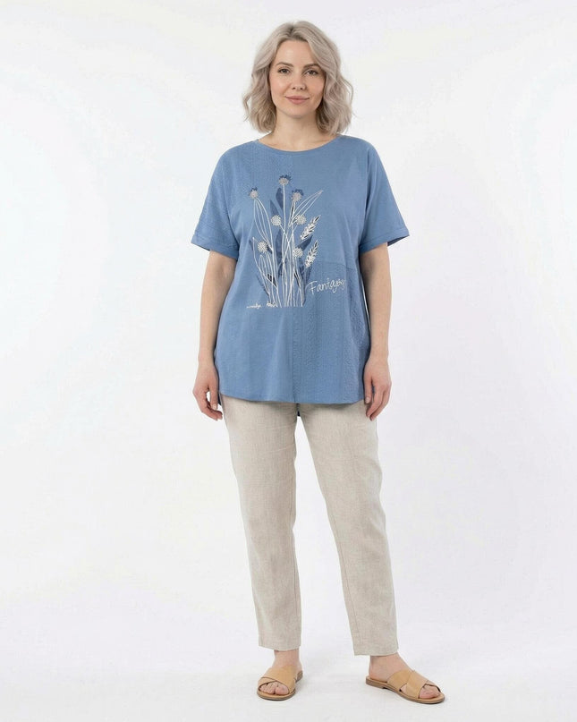 Ολόσωμη λήψη με μπλε καλοκαιρινή τουνίκ μεγάλα μεγέθη, διακοσμημένη με λευκά λουλούδια και συνδυασμένη με ανοιχτόχρωμο παντελόνι.