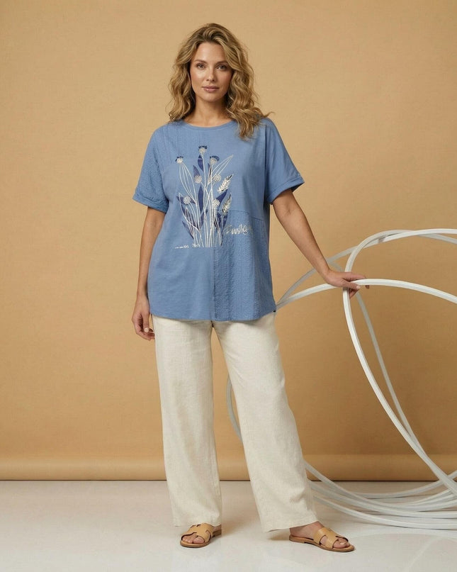 Μπλε καλοκαιρινή τουνίκ μεγάλα μεγέθη με φλοράλ απλικέ και δαντέλα. Ολόσωμη λήψη όπου το ρούχο συνδυάζεται με ένα λευκό παντελόνι.