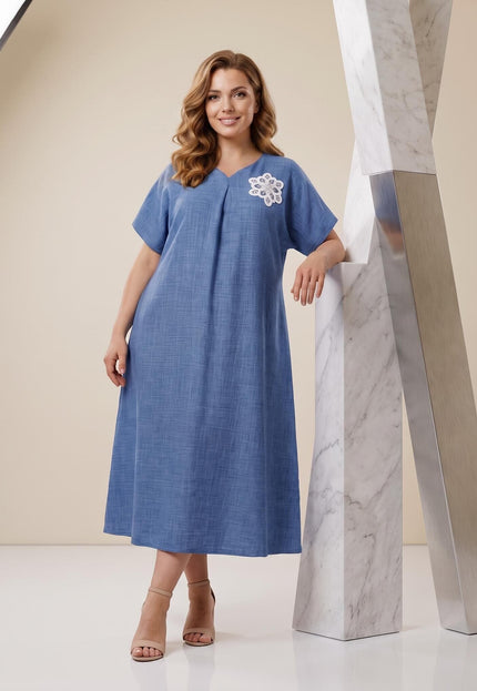 Ολόσωμη λήψη με κομψό μπλε μάξι φόρεμα plus size σε γραμμή Α. Ξεχωρίζει το V-ντεκολτέ, τα κοντά μανίκια και το λευκό φλοράλ κέντημα.
