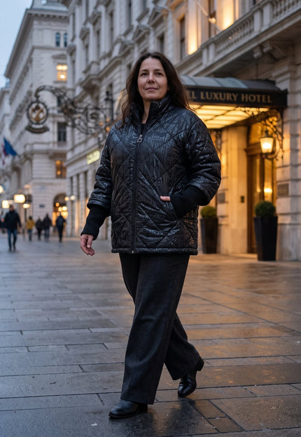 Γυναίκα φορά μαύρο καπιτονέ μπουφάν με μοτίβο γραμμάτων μπροστά σε φωτισμένη είσοδο ξενοδοχείου.